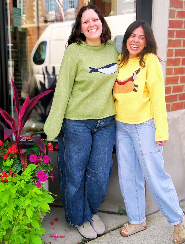 Two women standing outdoors, one in a green sweater with a fish design and the other in a yellow sweater with a cartoon character, in front of a brick building.